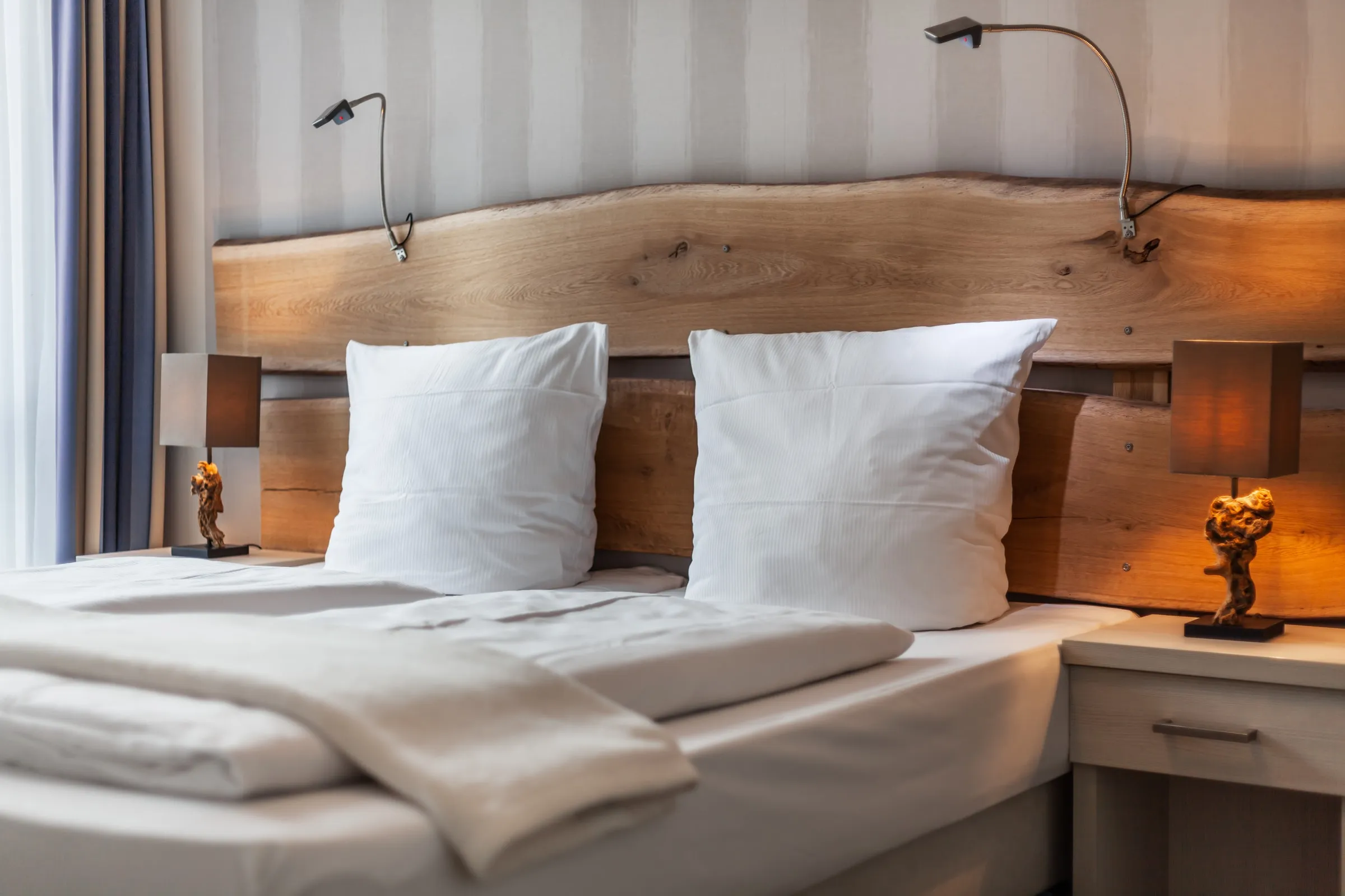 Das Doppelbett aus der Nähe: weiße Kissen, Kopfteil aus Holz, eine brennende Nachttischlampe