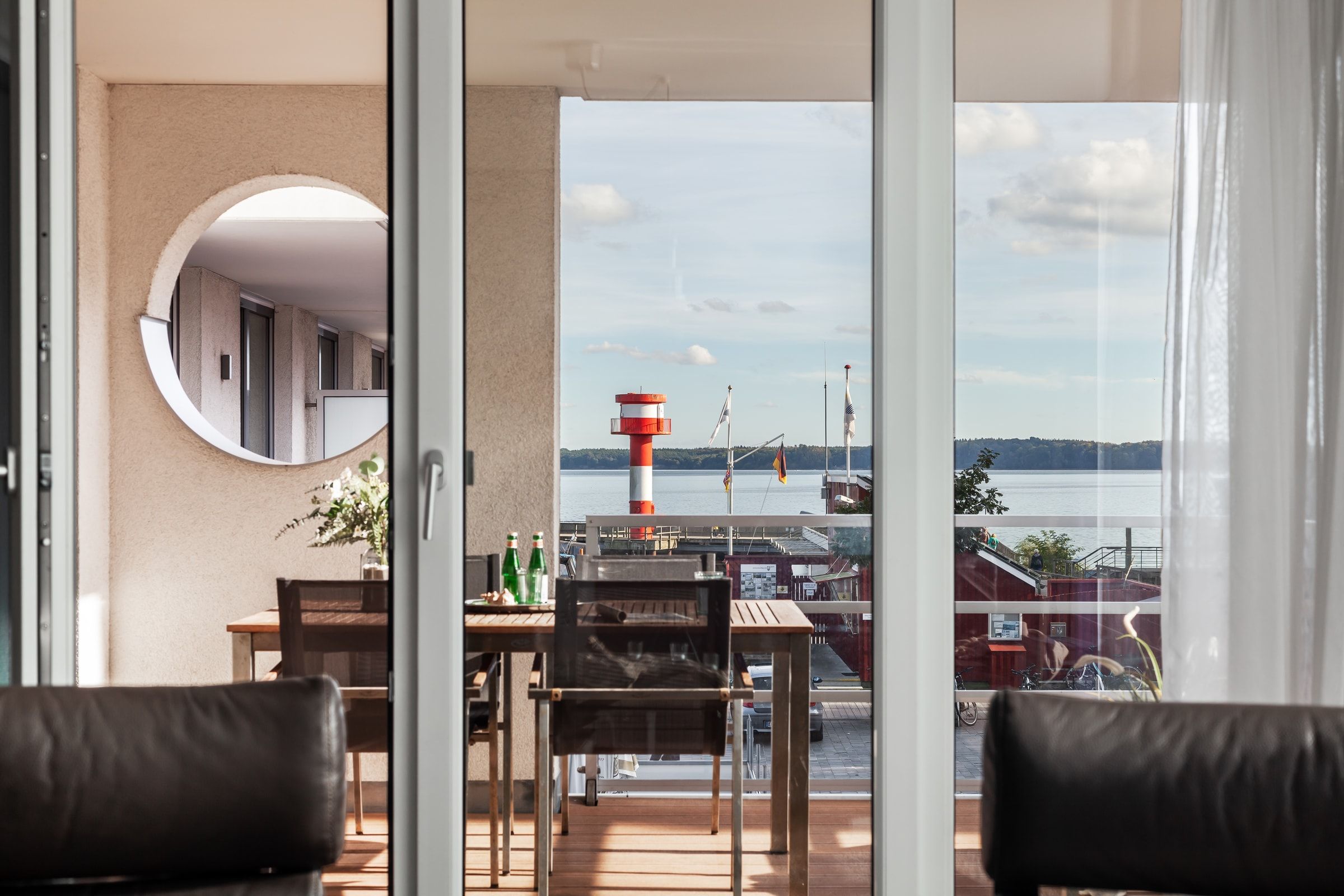 Blick aus dem Wohnraum durch die Glastür auf die Loggia: das runde Bullauge, der Tisch mit seinen Stühlen, dahinter der rot-weiße Leuchtturm und die Bucht