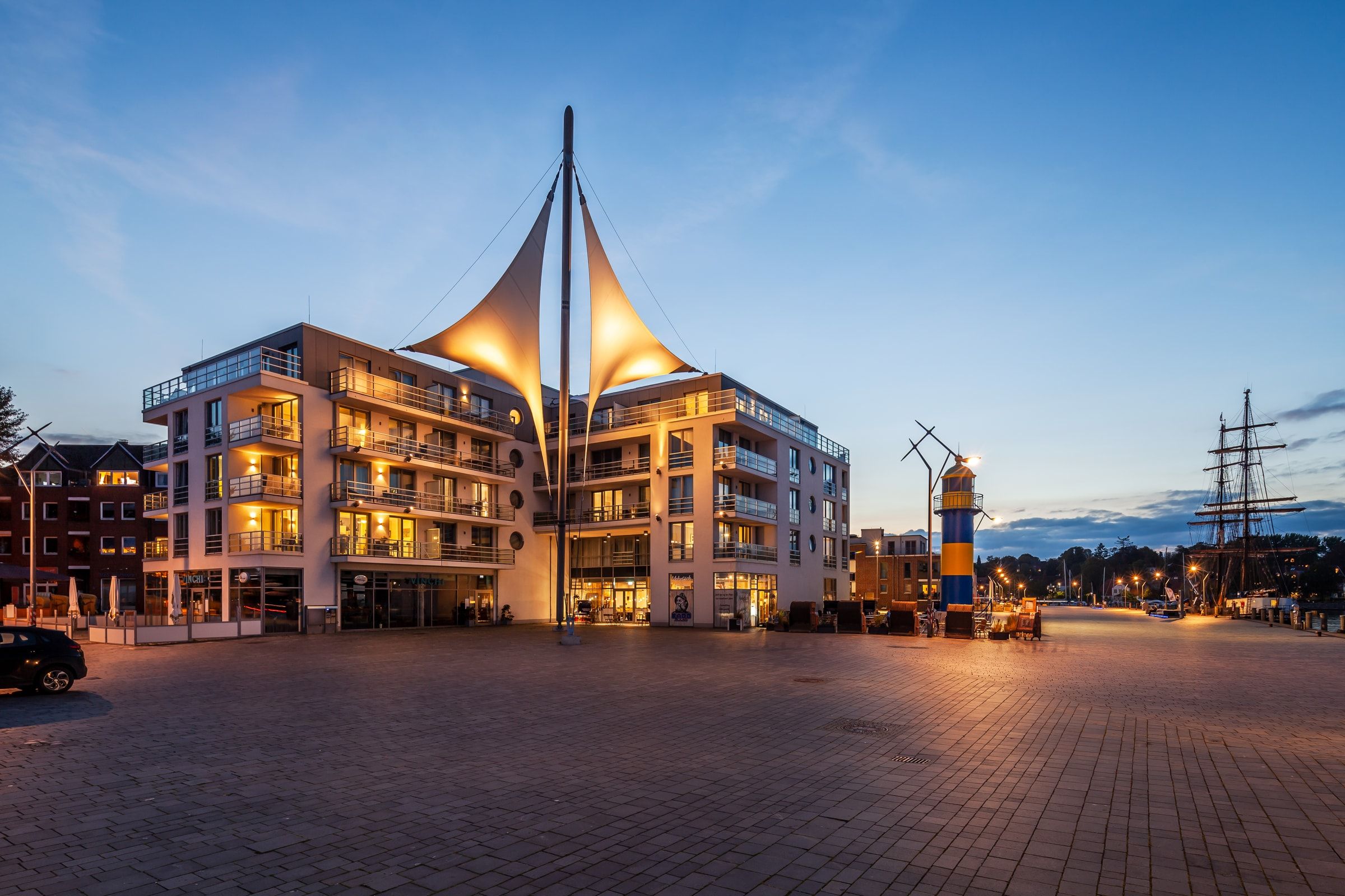 Das Apartmenthaus Hafenspitze am Abend, beleuchtete Loggien, rechts das weiße Segel über dem Hafenplatz