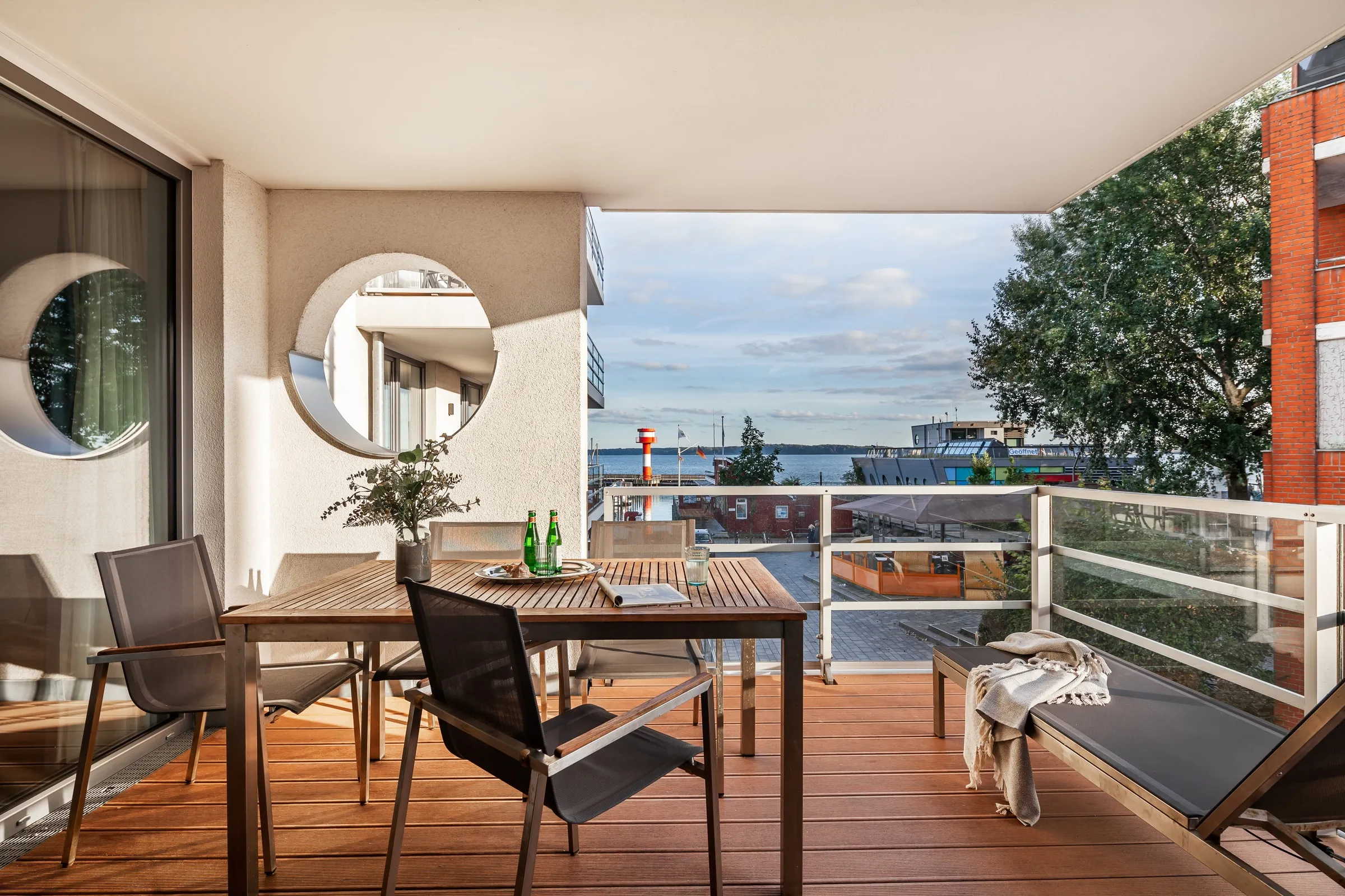 Die Loggia am Tag: Tisch und Stühle, das runde Bullauge, dahinter Hafen und Bucht